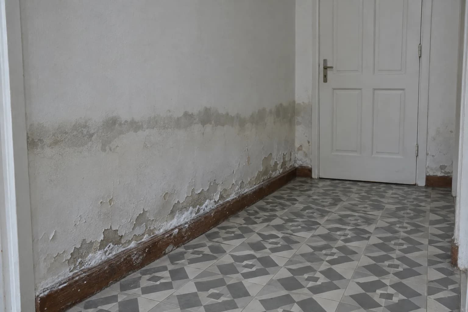 Parede de um corredor com tinta a descascar e sais brancos junto ao rodapé, sobre um piso de ladrilho hidráulico