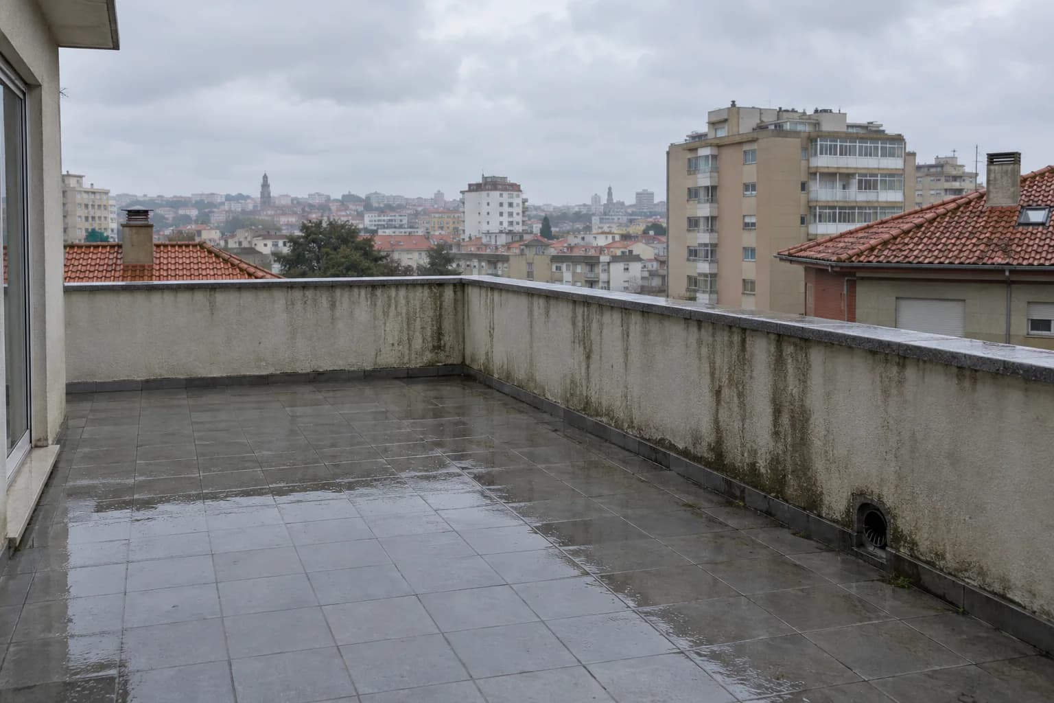 Terraço de um prédio depois da chuva, com o piso molhado e um muro de reboco manchado, sobre telhados da cidade